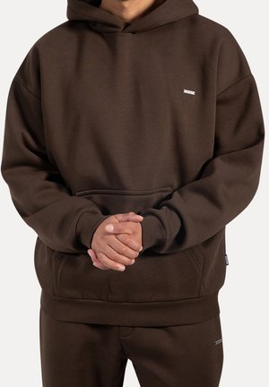 Person trägt einen dunkelbraunen Hoodie und passende Hose, Hände vor dem Körper gefaltet, schlichter Hintergrund.