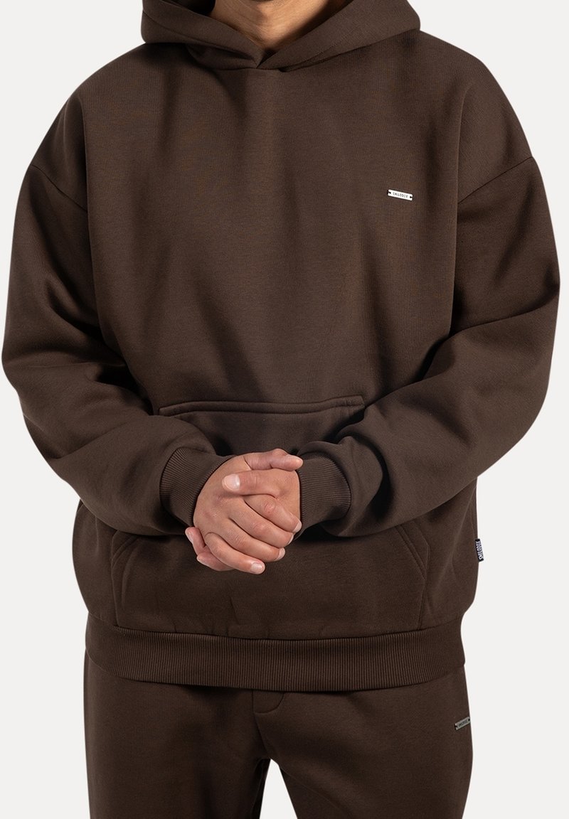 Personne portant un sweat à capuche marron foncé et un pantalon assorti, mains jointes devant, fond uni.