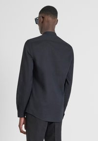 Camicia nera a maniche lunghe con colletto, dal design leggermente aderente, texture liscia, con dettagli di bottoni sui polsini. Indossata con pantaloni scuri.