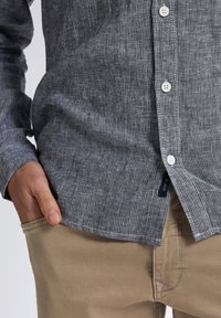 Camicia a bottoni a righe verticali blu scuro e grigie, con un tessuto testurizzato e bottoni bianchi. Abbinata a pantaloni beige, mano in tasca.