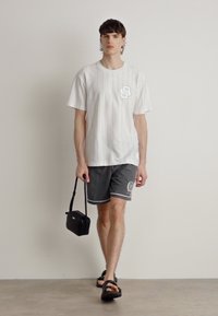 Hvit, kortermet skjorte med vertikale striper og en liten '8'-logo, paret med grå stripete shorts og svarte sandaler, holder en liten veske.