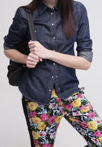 Donkerdenim overhemd met knopen aan de voorkant, borstzakken en opgerolde mouwen, gecombineerd met kleurrijke, bloemenpatronen broek met verschillende bloemen.