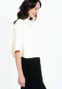Short-sleeve, cream blouse in lightweight fabric, featuring a button-down front and relaxed fit. Paired with a black skirt.
