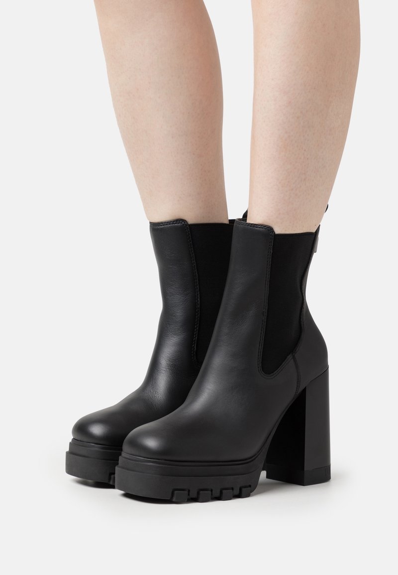 Bottes en cuir noir à la cheville avec talons hauts épais et semelles épaisses à crampons portées sur des jambes nues.