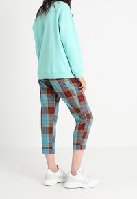 Persona che indossa un felpa azzurro chiaro, pantaloni a quadri colorati e scarpe bianche, in piedi di lato su uno sfondo semplice.