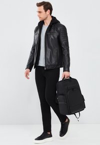 Chaqueta de cuero negra con acentos acolchados en los hombros, camiseta gris, pantalones negros y zapatillas negras. Lleva una mochila negra con múltiples bolsillos.