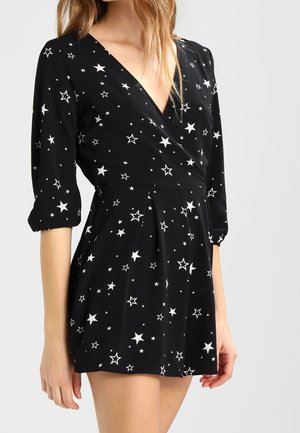 Czarna sukienka kopertowa z białymi wzorami gwiazd, z rękawami do połowy przedramienia i plisowaną spódnicą, noszona przez kobietę o jasnej karnacji i blond włosach.