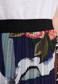 Fascia elastica nera con dettaglio testurizzato; il tessuto plissettato presenta motivi floreali e astratti in tonalità di blu navy, verde, pink e beige.