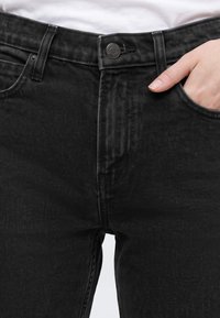 Gros plan d'une personne portant un jean noir avec la main droite partiellement dans la poche avant, ceinture et bouton visibles.