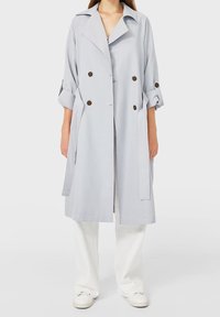 Osoba w długim, jasnoszarym płaszczu z podwiniętymi rękawami, z brązowymi guzikami, białych spodniach i białych trampkach na jednolitym tle.