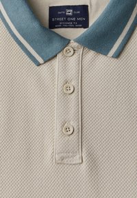 Vit pikétröja med texturerad tyg, turkos krage med vita ränder och knäppning med tre knappar. Varumärkesetikett synlig.