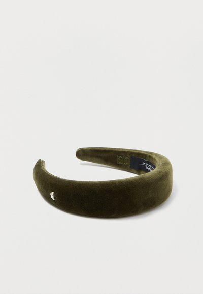 Diadema de terciopelo verde oliva con una textura suave, forma redondeada y pequeño detalle del logo en blanco en el lateral. Diseño ligero y flexible.