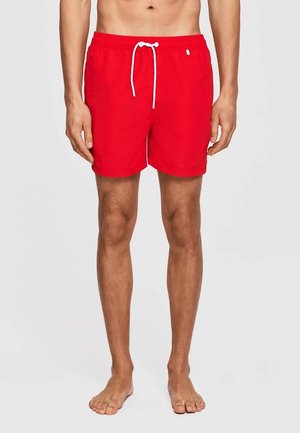 Mężczyzna ubrany w proste czerwone kąpielówki ze białym regulowanym sznurkiem, stojący boso na neutralnym szarym tle.