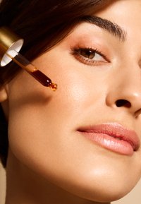 Ansigtserumdråber med ravfarvet væske påføres en glat, strålende kind. Hudtonen er lys med en subtil glans og naturlig makeup.