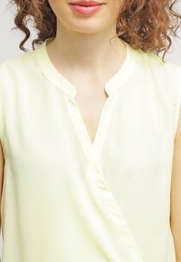 Light yellow blouse with a V-neckline, short sleeves, and a smooth, soft texture. Features a wrap design and subtle stitching details.