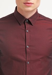 Burgundská košile na knoflíky se strukturovaným límcem, s hladkou texturou a pěti šedými knoflíky vpředu.