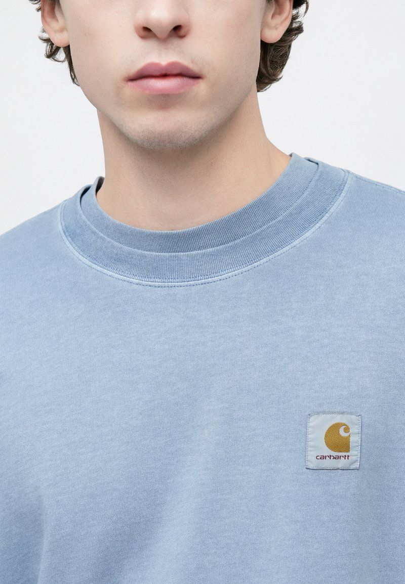 Sweat-shirt à col rond bleu clair en tissu doux. Présente un patch logo carré jaune et blanc sur le côté gauche de la poitrine.