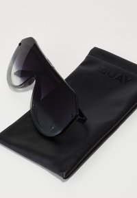 Juodos didelio dydžio saulės akinės su gradientiniais pilkais stiklais, turinčios elegantišką dizainą ir kampinį rėmą, guli ant juodos odinės dėklo, išraižyto žodžiu "QUAY."