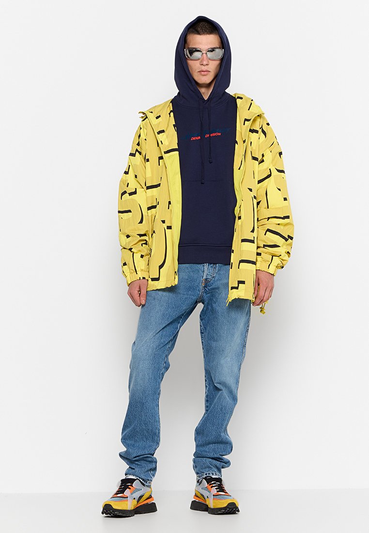 Veste jaune avec des motifs abstraits noirs, sweat à capuche bleu marine, jeans bleus et baskets multicolores. La veste a des poignets élastiques et une fermeture éclair.