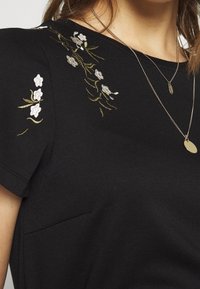 Top corto nero a maniche corte con complessa ricamo floreale in bianco e verde lungo la spalla, con scollatura rotonda e dettaglio della tasca.