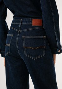 Mörkblå denimjeans med rak skärning, med en brun läderlapp på baksidan, gula sömmar och två bakfickor.