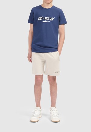 Marineblaues T-Shirt mit weißem Grafikdesign, kombiniert mit beigen Shorts mit schwarzem Logo. Die Schuhe sind weiß mit strukturiertem Stoff und Kautschuksohle.