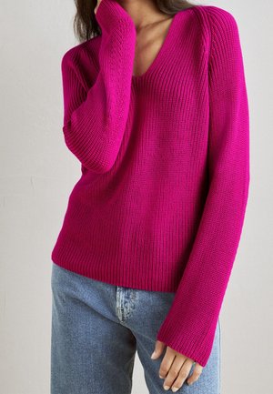 Woman wearing a bright pink ribbed knit sweater with a V-neck and light blue jeans, one hand touching her face.