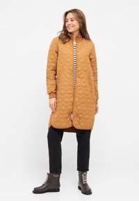 Vadderad brun lång jacka med ribbad krage, dragkedja, sidoknappar, bärs över en randig skjorta och matchas med grå snörkängor.