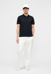 Fiatal férfi, aki zsebre tett kézzel áll, fekete pólóinget, fehér nadrágot, fehér sportcipőt és fehér baseballsapkát visel.