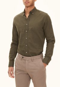 Olijfgroene overhemd met lange mouwen, gestructureerde stof, contrasterende knopen, gedragen met lichtbeige broeken, handen in de zakken.