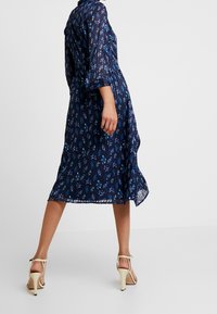 Námořnická modrá květinová šaty s tříčtvrtečními rukávy, splývavým materiálem, délkou ke kolenům a volánkovým lemem. V kombinaci se světlými vysokými sandály.