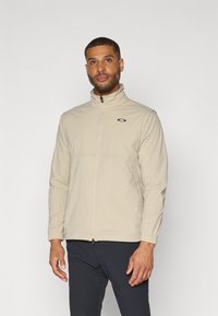 Veste zippée beige avec col haut, en matériau léger, avec des poignets élastiques et un logo brodé sur la partie gauche de la poitrine.