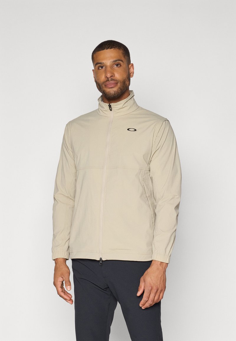 Veste zippée beige avec col haut, en matériau léger, avec des poignets élastiques et un logo brodé sur la partie gauche de la poitrine.