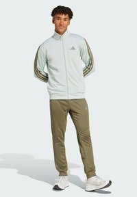 Ljusblå zip-up jacka med bruna ränder och logotyp, tillsammans med olivgröna joggingsbyxor. Vita sportskor fulländar outfiten.