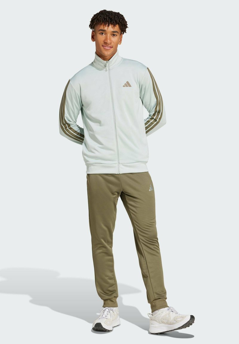 Ljusblå zip-up jacka med bruna ränder och logotyp, tillsammans med olivgröna joggingsbyxor. Vita sportskor fulländar outfiten.