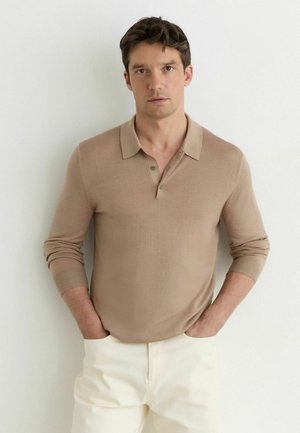 Man met donker haar die een beige poloshirt met lange mouwen en een witte broek draagt, staand met handen in de zakken tegen een witte achtergrond.