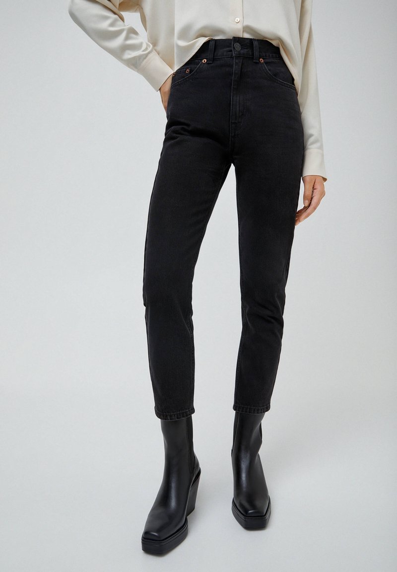 Personne portant un jean noir taille haute avec poches et des bottines noires à talons, associée à une chemise crème à manches longues rentrée dedans.