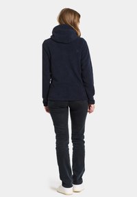 Marineblauer Fleece-Hoodie mit hohem Kragen, strukturiertem Stoff und langen Ärmeln, kombiniert mit schwarzen Hosen und weißen Sneakers.