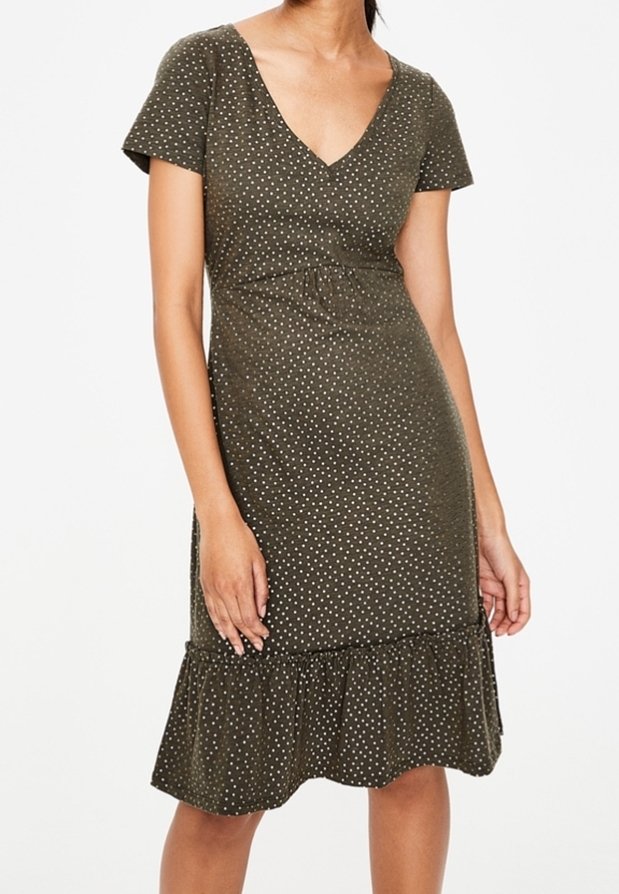 Femme portant une robe courte manches courtes vert olive à pois blancs, col en V et ourlet volanté, debout devant un fond uni.