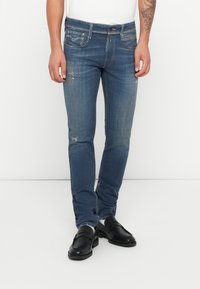 Jeans de mezclilla azul de corte slim con ligeros desgastes y detalles desgatados, con cinco bolsillos y cierre de botón. Combinados con mocasines negros.
