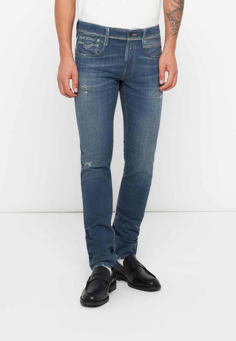 Jeans de mezclilla azul de corte slim con ligeros desgastes y detalles desgatados, con cinco bolsillos y cierre de botón. Combinados con mocasines negros.