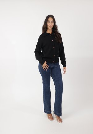 Cardigan noir en tricot avec un design à col et des boutons marron, associé à un jean évasé bleu et des sandales transparentes. Look simple et classique.