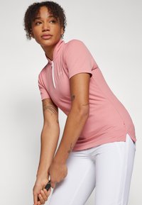 Ροζ πόλο μπλούζα με κοντά μανίκια, υψηλό κολάρο και λευκό φερμουάρ. Διαθέτει ριγέ υφή και λογότυπο CK. Συνδυάζεται με λευκά παντελόνια.