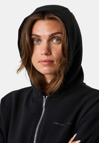 Schwarze Zip-Hoodie mit gefütterter Kapuze, ausgestattet mit einem silbernen Reißverschluss und einem kleinen Logo auf der Brust. Glatter Stoff mit lockerer Passform.