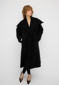 Cappotto di lana nero con colletto ampio, tasche e chiusura a un bottone. Abbinato a leggings neri e scarpe a punta.