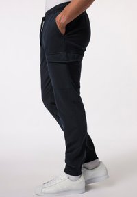 Man in zwarte cargo joggers met enkelmanchetten en witte sneakers, met een hand nonchalant in de zak, staand tegen een effen achtergrond.