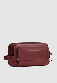 Toilettas van bordeauxrood leer met een soepel oppervlak, dubbele ritssluitingen en een zijhandvat voor gemakkelijk dragen. Zilveren logodetail zichtbaar.
