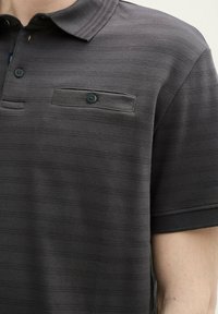 Ciemnoszara koszulka polo z teksturowanym, paskowanym wzorem. Posiada klasyczny kołnierzyk, dwie kieszenie na piersi oraz akcenty w postaci guzików.
