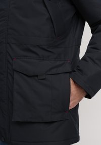 Black padded jacket featuring a large front pocket with a flap, subtle red accents, and a smooth, textured fabric.