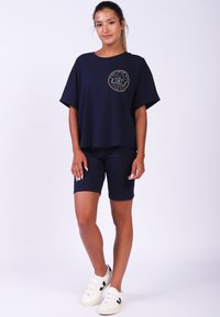 Navyblauw T-shirt met korte mouwen en een ronde grafische embleem, gecombineerd met navy shorts. Witte sneakers met drie banden en zwarte accenten.